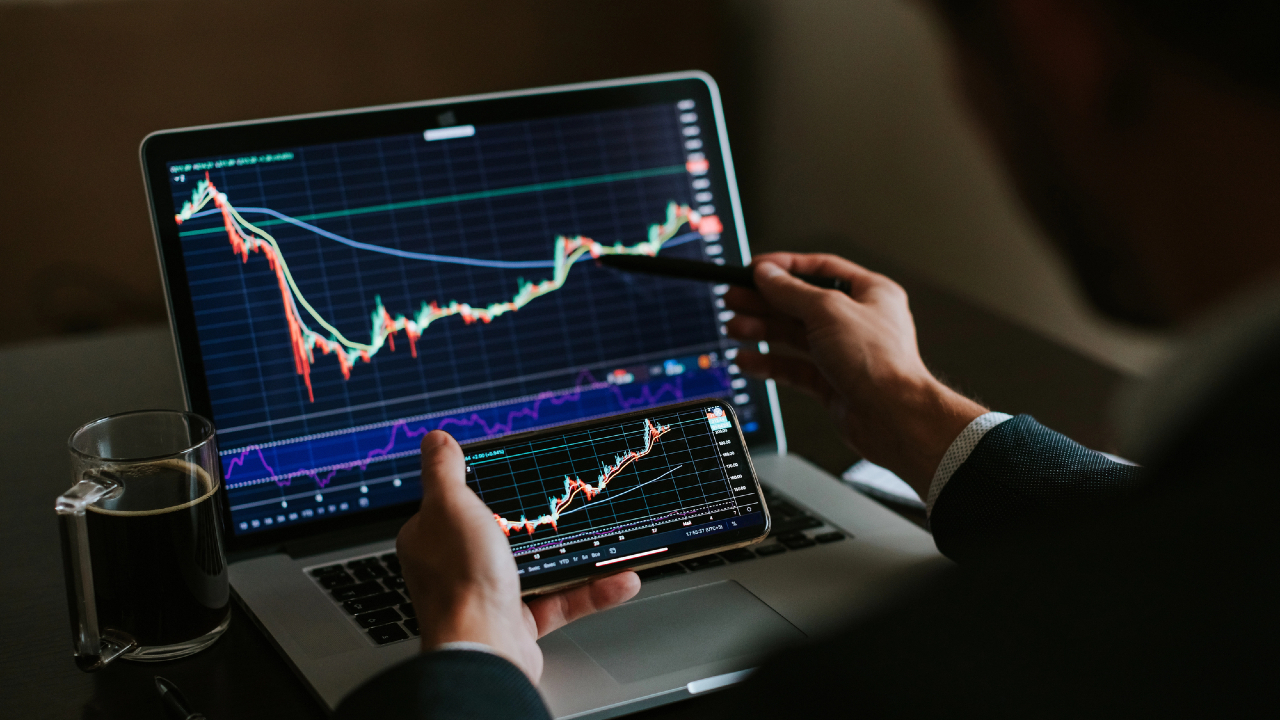 Una persona analizza grafici finanziari su un laptop e un tablet, esaminando attentamente grafici lineari con tendenze fluttuanti, forse correlate ai titoli alto dividendo nei mercati azionari. Una penna punta verso lo schermo mentre una tazza di caffè è lì vicino.