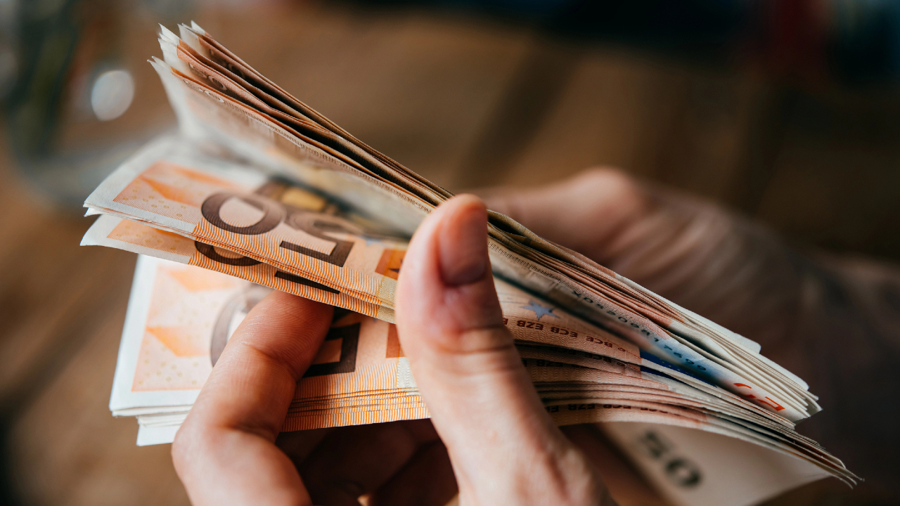 Una persona conta meticolosamente una pila di banconote in euro, forse pensando a come spendere la loro tredicesima. L'immagine si ingrandisce sulle mani e sulle banconote vibranti, su uno sfondo leggermente sfocato.