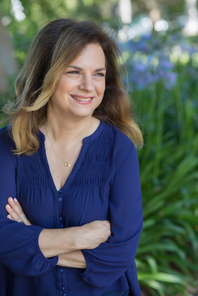 Una donna con i capelli castani lunghi fino alle spalle, che indossa una camicetta blu, è in piedi con le braccia incrociate. Sta sorridendo e guarda di lato. Lo sfondo presenta fogliame verde e accenni di fiori viola.