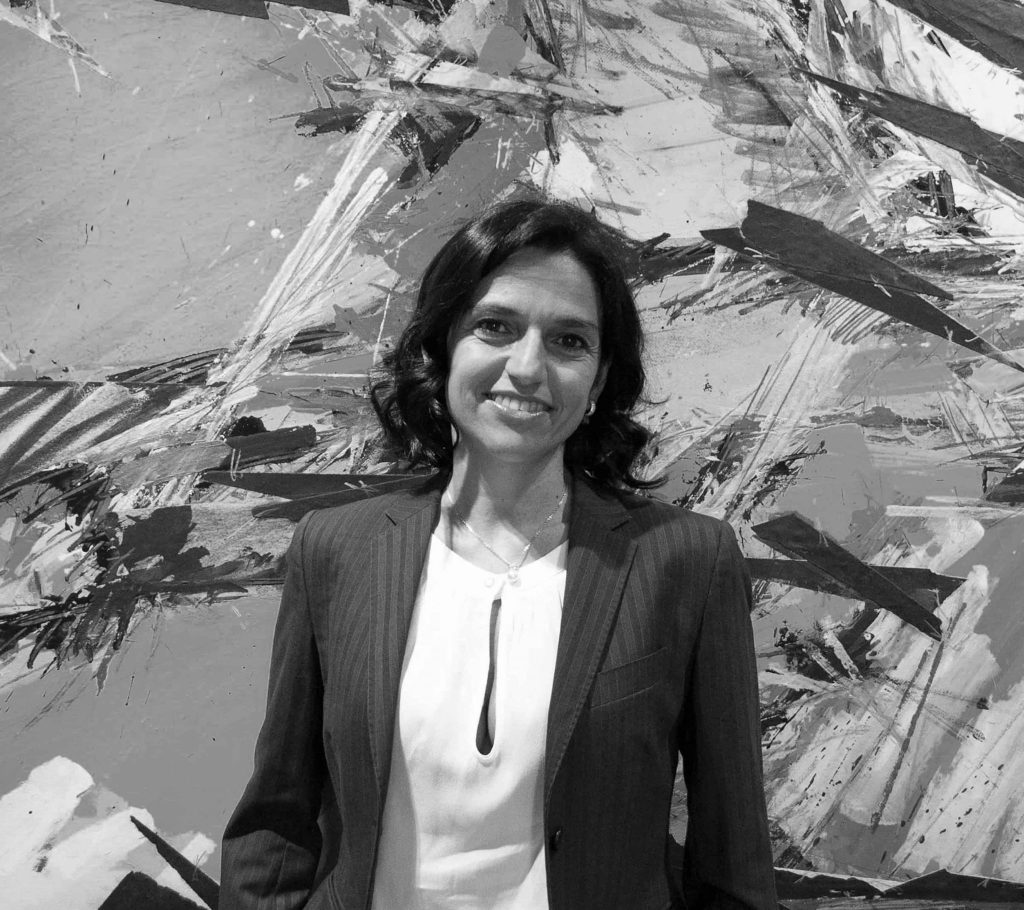 Una donna con i capelli scuri lunghi fino alle spalle, che indossa un blazer e una camicetta chiara, sorride in piedi davanti a uno sfondo astratto dipinto in bianco e nero.