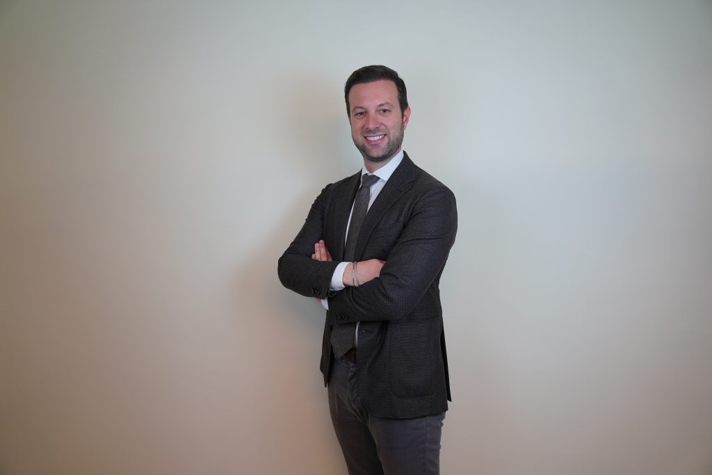 Un uomo in abito scuro e cravatta è in piedi su uno sfondo chiaro e semplice, sorridente e con le braccia incrociate.