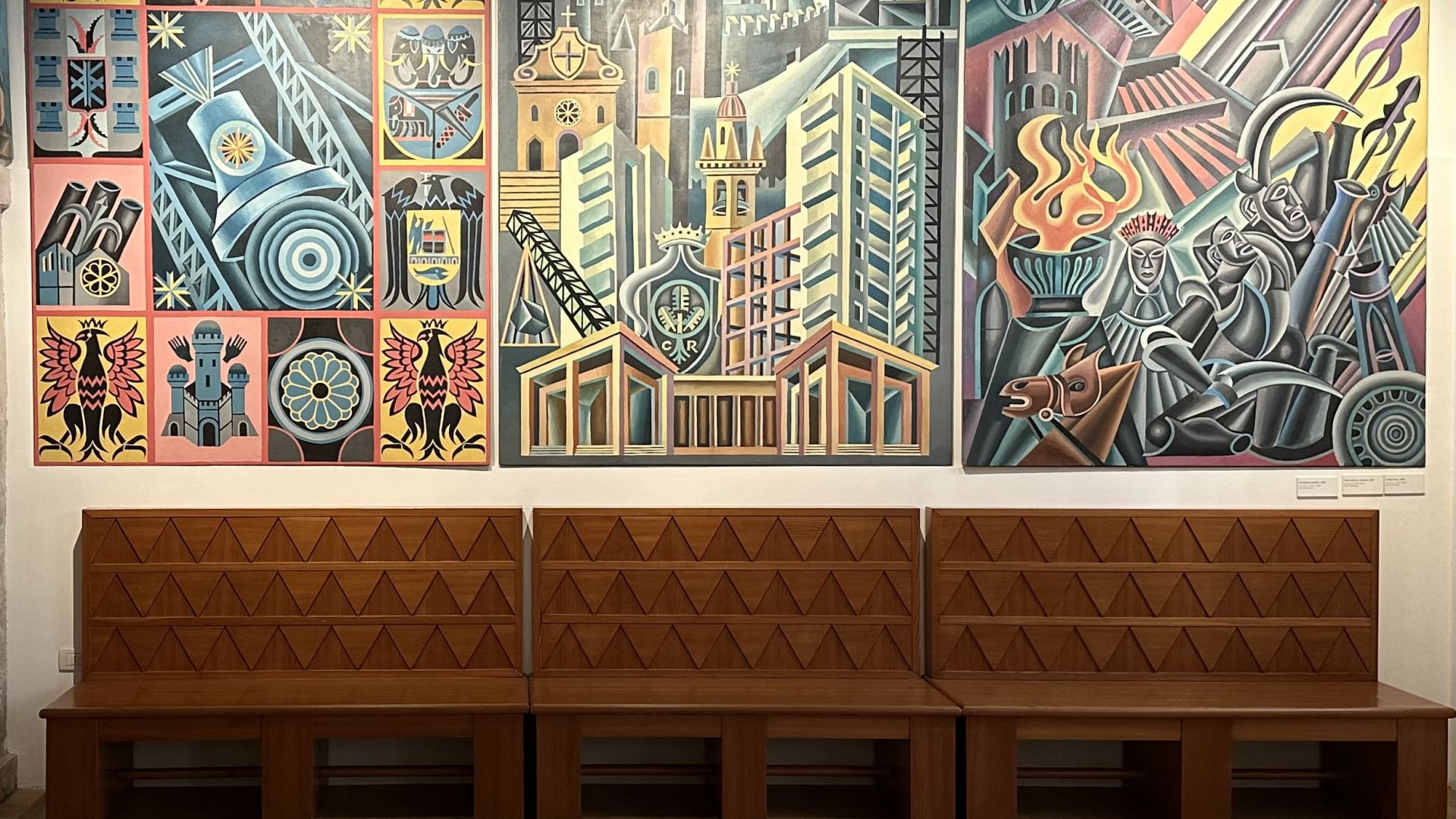 Panche di legno di fronte a tre murales colorati ispirati a Depero Rovereto, raffiguranti paesaggi urbani astratti e geometrici e scene dinamiche con elementi architettonici e motivi vivaci. Le composizioni includono edifici, ponti e figure stilizzate.