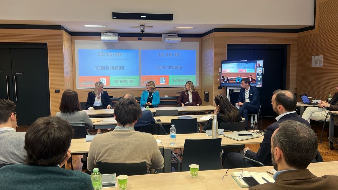 Un gruppo di persone sta partecipando a una tavola rotonda in una sala conferenze. Quattro relatori sono seduti a un tavolo di fronte al pubblico. La sala ha due schermi che mostrano "CO-WIN" e i partecipanti guardano attentamente, seduti con computer portatili e documenti.