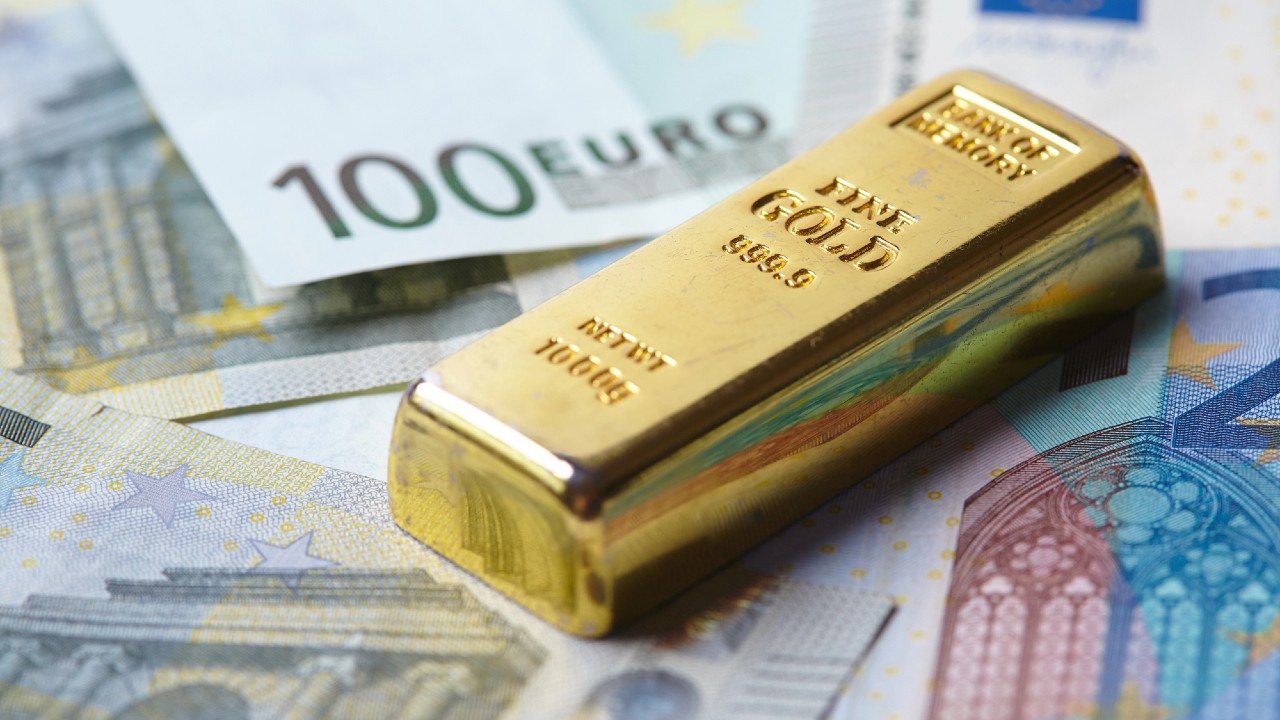 Una barra d'oro è appoggiata su banconote da 100 euro sparse. La barra è incisa con dettagli come purezza "999,9" e "NET WT 1000g". La scena riflette i temi della ricchezza e degli investimenti.