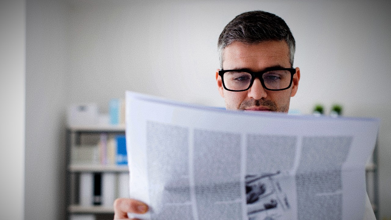 Una persona con gli occhiali legge un giornale in un ambiente chiuso. Lo sfondo è sfocato, e mostra scaffali con libri e oggetti decorativi. L'attenzione è sul volto e sul giornale.