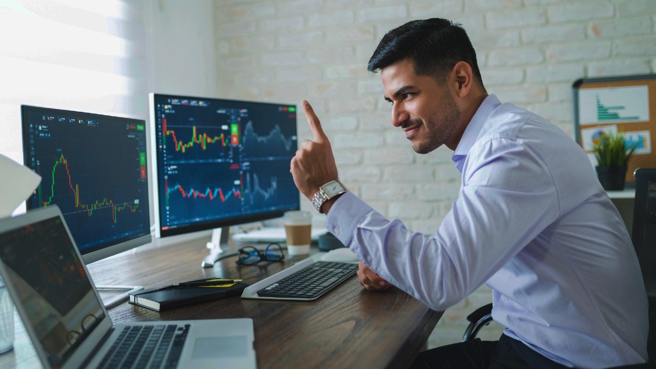 Un uomo in camicia bianca è seduto a una scrivania, analizza i dati su più monitor di computer che mostrano grafici e diagrammi. Tiene un dito alzato, con un'espressione concentrata. Forniture per ufficio e una pianta sono visibili sulla scrivania.