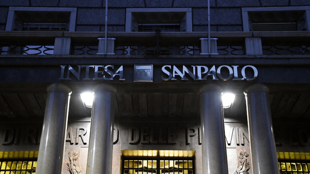 L'immagine mostra la facciata di un edificio con la scritta "Intesa Sanpaolo" illuminata di notte. Quattro grandi colonne sostengono la struttura e diverse luci illuminano l'area d'ingresso.