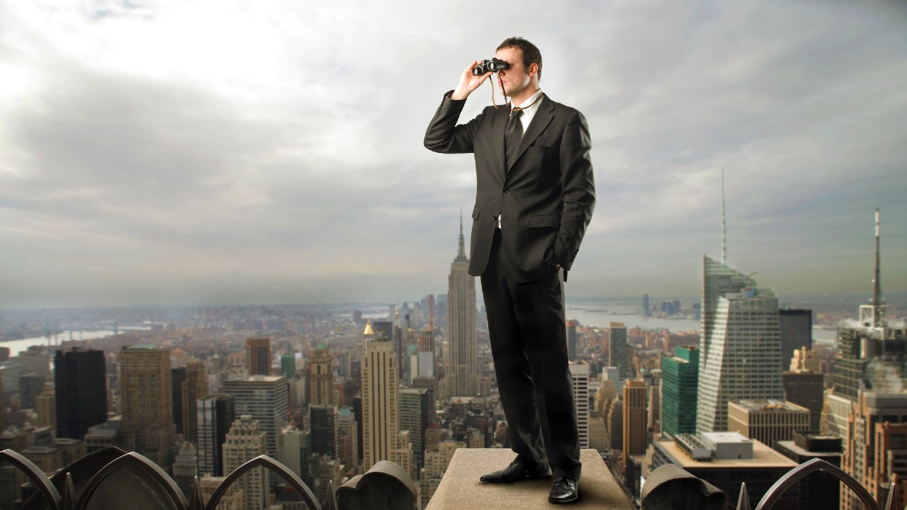 Un uomo in giacca e cravatta è in piedi su un'alta sporgenza che domina un paesaggio urbano. Sta usando un binocolo per guardare in lontananza. Lo skyline della città, compresi gli alti edifici, è visibile sotto un cielo nuvoloso.