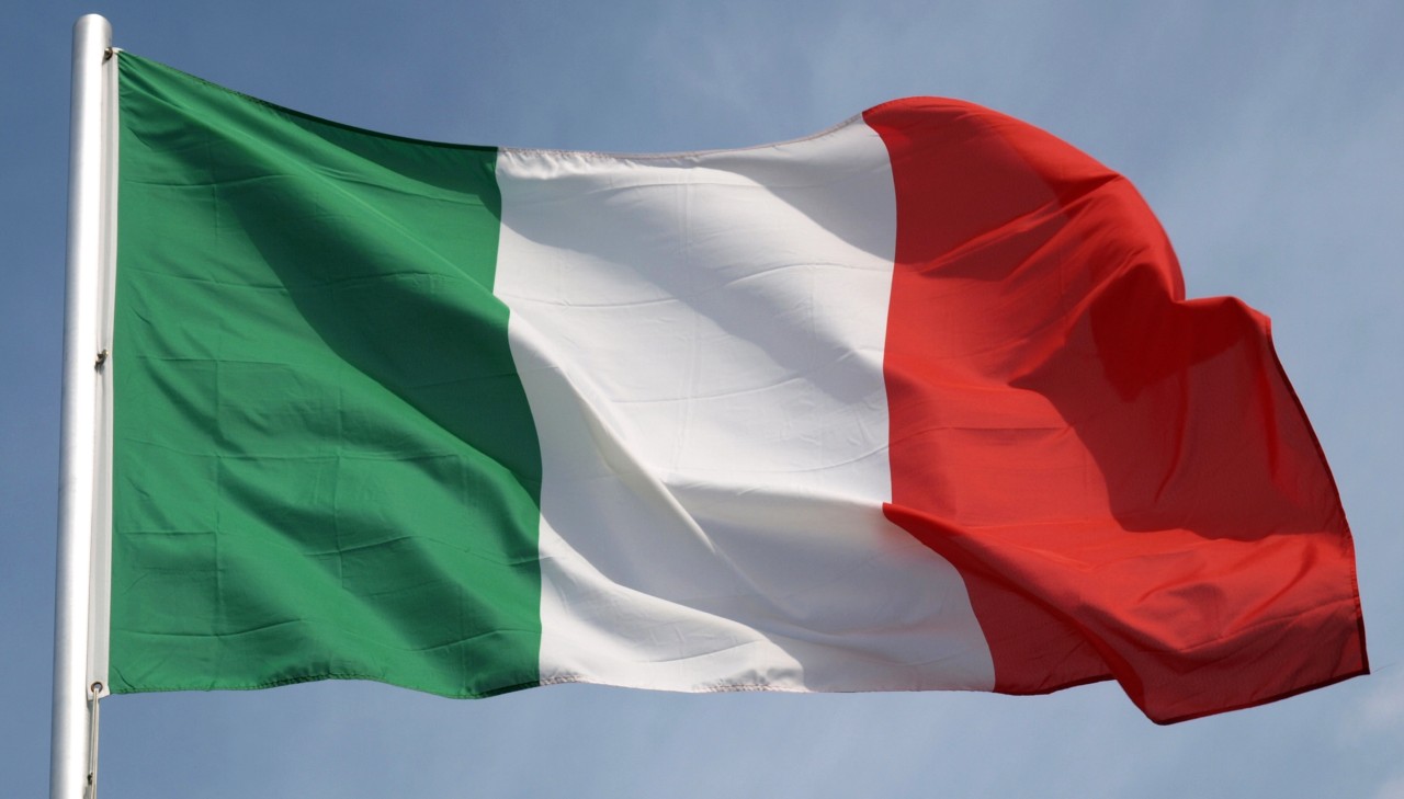 L'immagine mostra la bandiera nazionale dell'Italia che sventola al vento contro un cielo limpido. La bandiera è composta da tre strisce verticali: verde a sinistra, bianca al centro e rossa a destra.