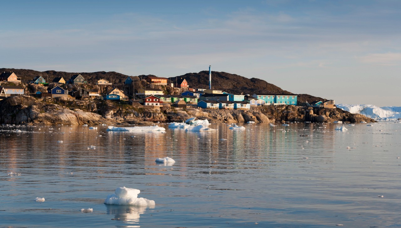 Un piccolo insediamento costiero con case colorate si trova su un terreno roccioso accanto a un mare calmo. Gli iceberg galleggiano nell'acqua sotto un cielo azzurro e limpido. Le colline accidentate formano lo sfondo.
