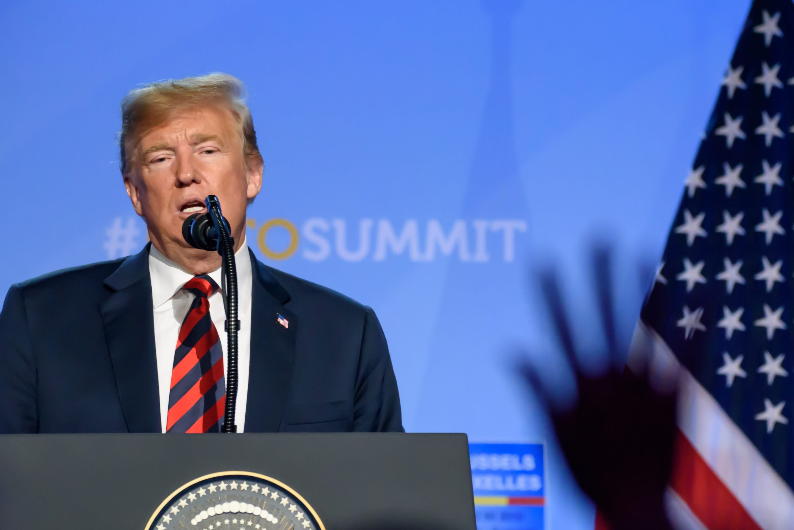 Un uomo parla su un podio con un microfono. Sullo sfondo c'è il testo "#NATOSUMMIT" e un'immagine sfocata della bandiera americana. Una mano è alzata in primo piano.