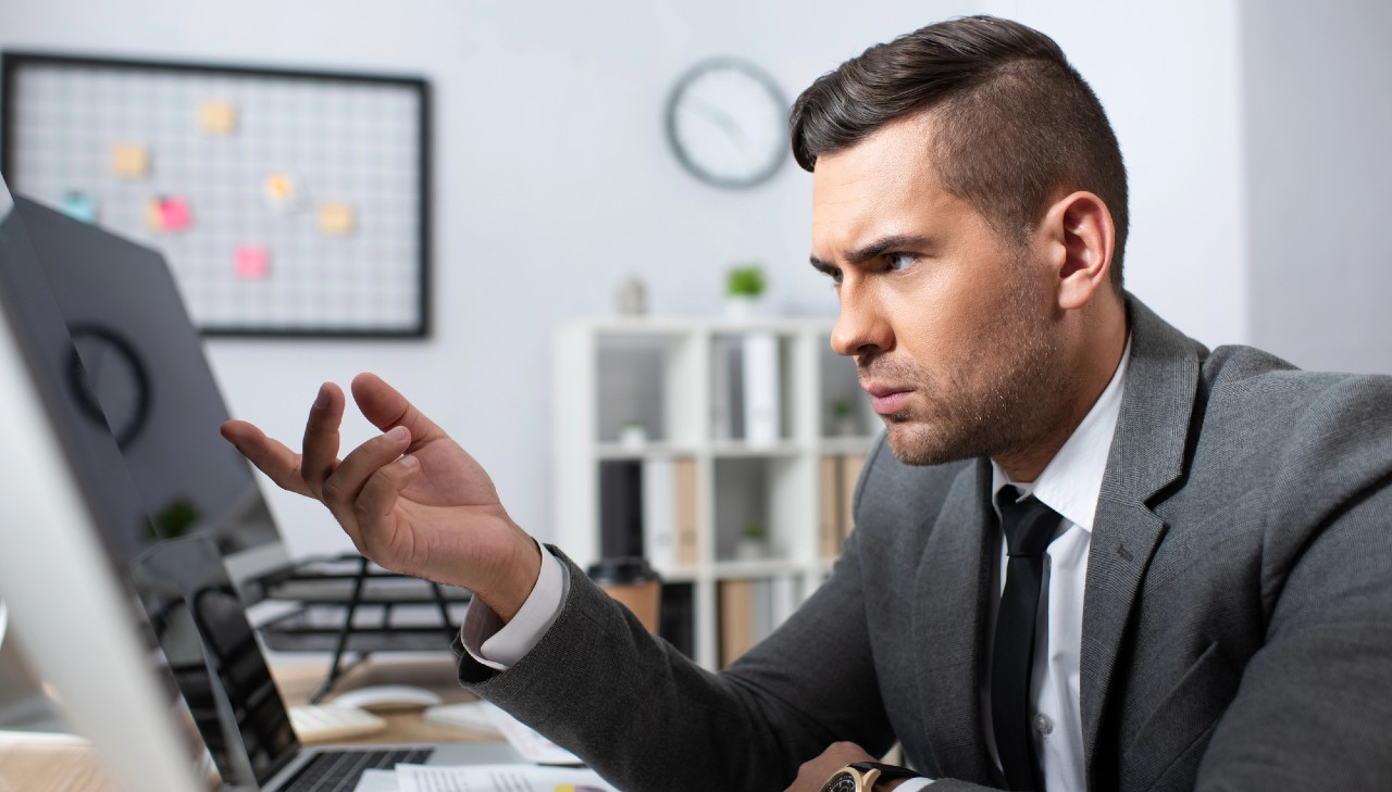 Un uomo in giacca e cravatta è seduto a una scrivania, guardando intensamente lo schermo di un computer. La sua mano è alzata con un gesto interrogativo. Lo sfondo include un ambiente d'ufficio sfocato con uno scaffale, un orologio e una bacheca di sughero con post-it.