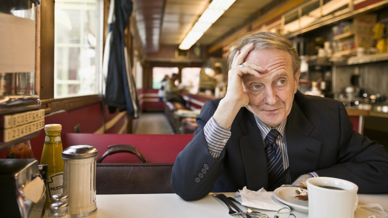 Un uomo anziano in giacca e cravatta siede in un tavolo da pranzo, appoggiando la testa sulla mano. Sembra pensieroso. Il tavolo ha una tazza da caffè, un piattino e un porta tovaglioli. Il ristorante ha un interno retrò con tavoli rossi e un lungo bancone.