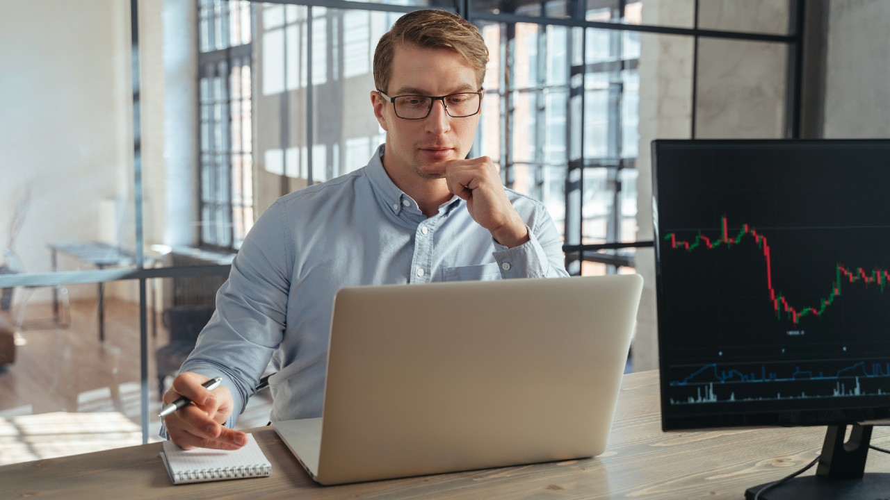 Un uomo con gli occhiali, in una camicia azzurra, è seduto a una scrivania in un ufficio moderno. È concentrato su un portatile, tiene una penna e un quaderno accanto a sé. Un monitor mostra un grafico a linee con dati finanziari. Grandi finestre sono sullo sfondo.