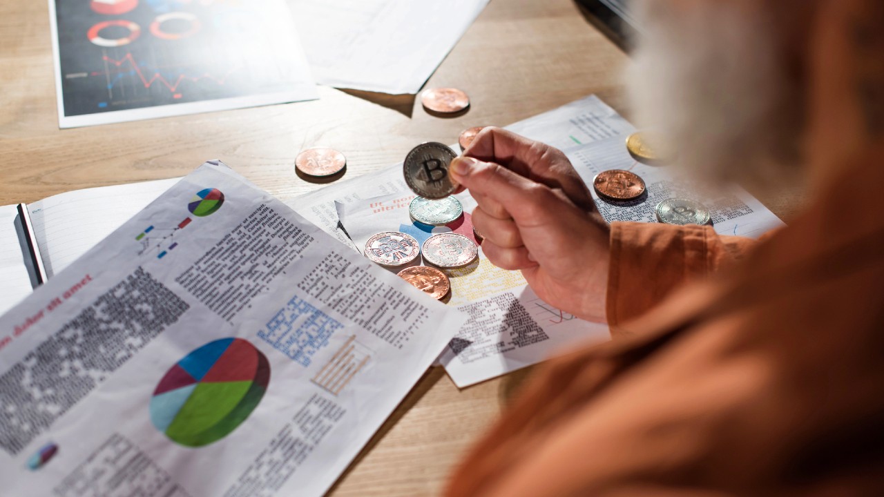 Una persona che tiene in mano una moneta Bitcoin in mezzo a giornali finanziari aperti e grafici su una scrivania. La scena include varie criptovalute, grafici a torta e grafici, che suggeriscono analisi e investimenti in valute digitali.