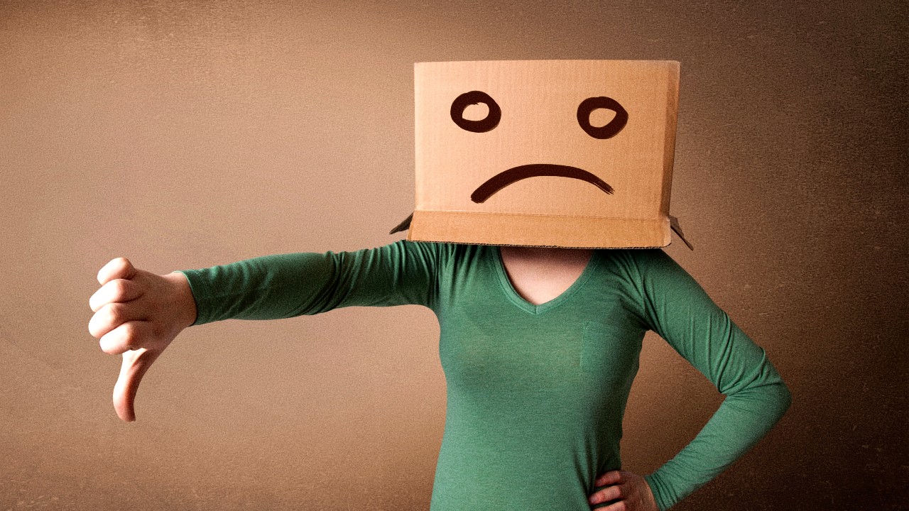 Una persona che indossa una maglietta verde è in piedi con una scatola di cartone sopra la testa. Sulla scatola è disegnata una faccia accigliata. La persona sta facendo un gesto di pollice verso con una mano. Lo sfondo è semplice e beige.