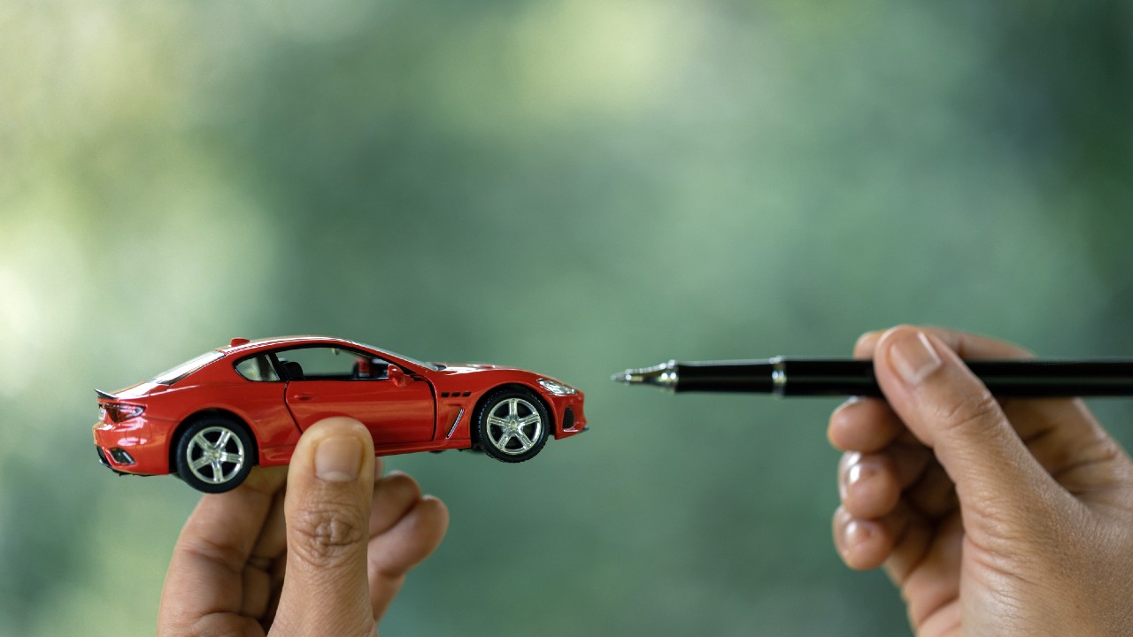 Una mano tiene una piccola macchinina rossa giocattolo di fronte a un'altra mano che tiene una penna, su uno sfondo verde sfocato. La penna è puntata verso la macchinina, creando una giocosa giustapposizione di scala.