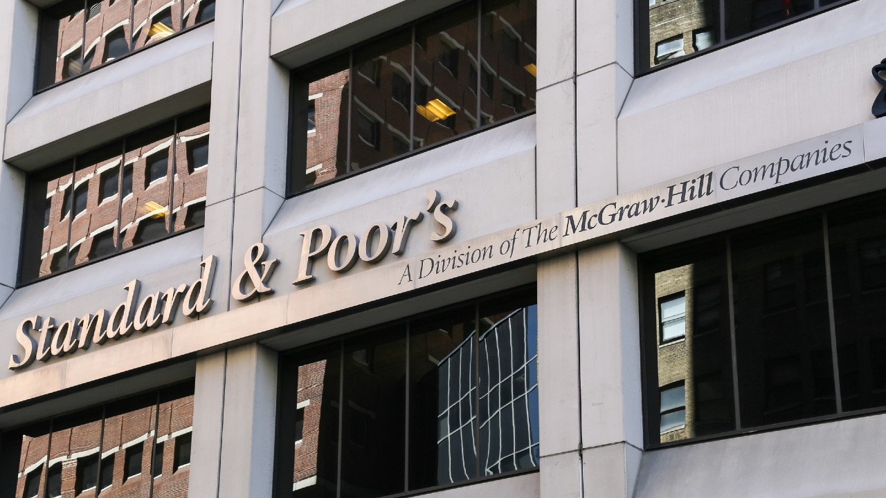 La facciata di un edificio con il cartello "Standard & Poor's, A Division of The McGraw-Hill Companies". La struttura è in cemento con finestre che riflettono gli edifici circostanti.