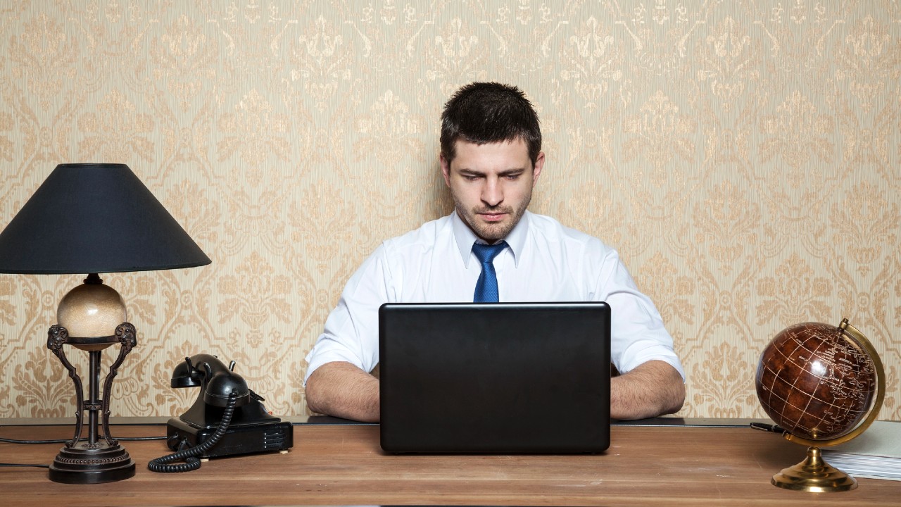 Uomo in camicia bianca e cravatta blu che lavora su un computer portatile a una scrivania di legno. Un telefono nero vintage, una lampada e un mappamondo sono appoggiati sulla scrivania. Lo sfondo è una carta da parati a motivi.