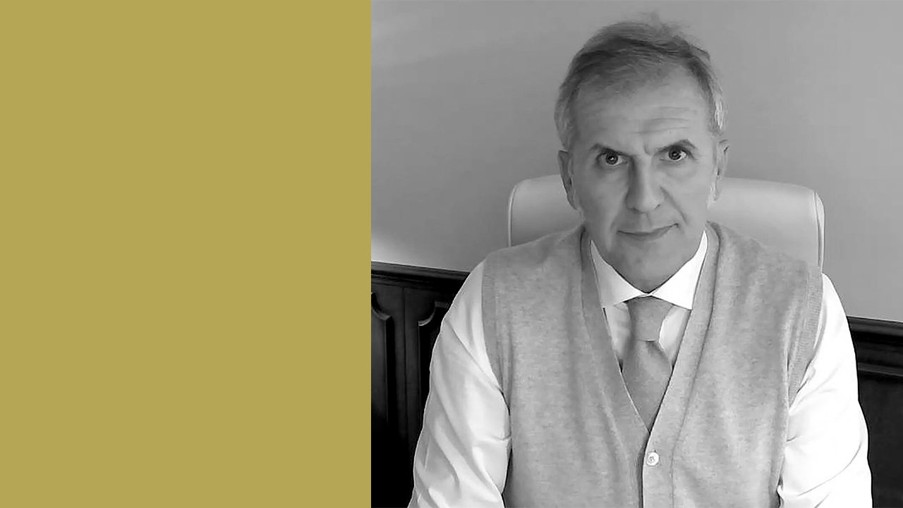 Foto in bianco e nero di una persona seduta su una sedia da ufficio, che indossa un maglione leggero sopra una camicia bianca e una cravatta. L'espressione della persona è neutra. Il lato sinistro dell'immagine ha uno sfondo giallo senape.