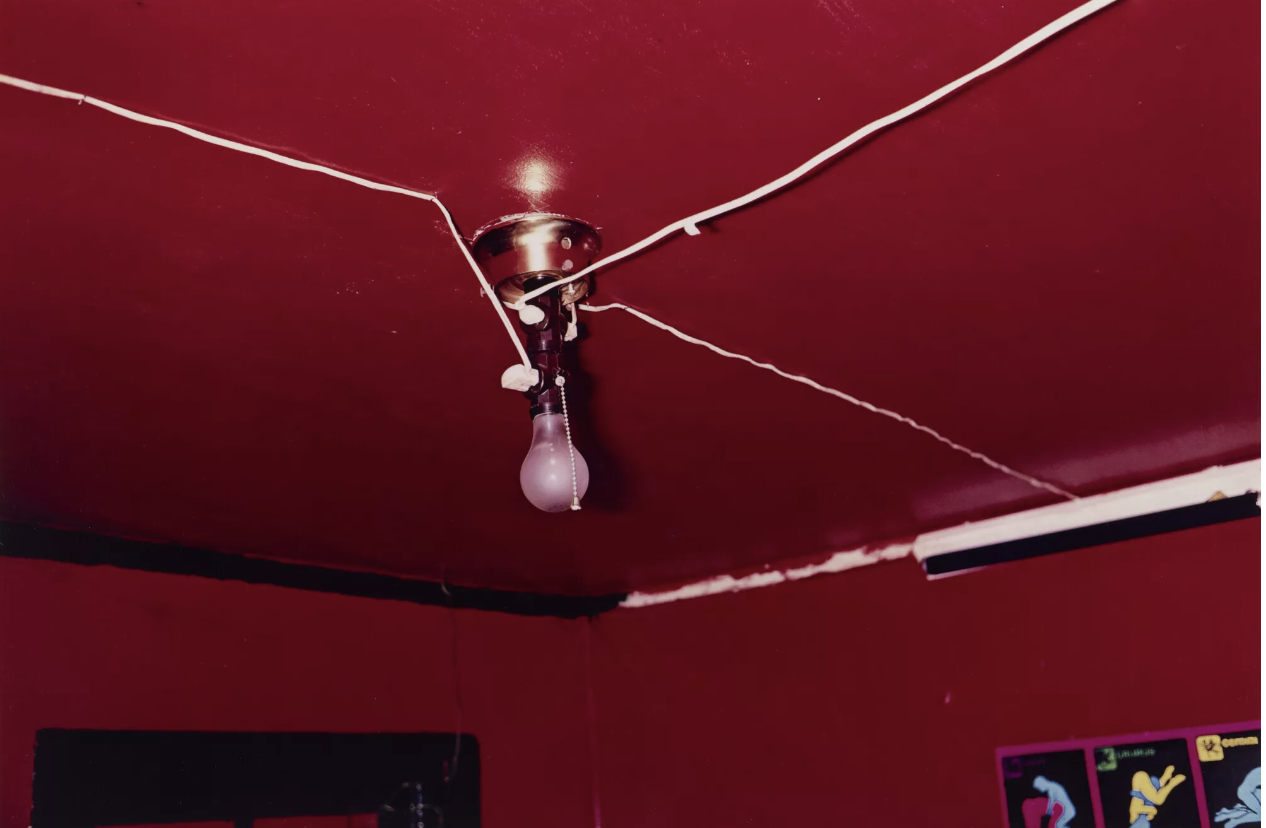 Una lampadina nuda pende da un soffitto rosso con fili esposti. Anche le pareti della stanza sono rosse. Nell'angolo è visibile parte di un poster con una grafica colorata. Il soffitto sembra leggermente invecchiato, con la vernice scrostata in alcune aree.