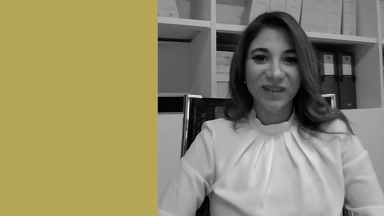Una donna con i capelli lunghi e una camicetta chiara è seduta su una sedia da ufficio. Dietro di lei ci sono scaffali con raccoglitori e scatole. Il lato sinistro dell'immagine è di un solido color senape. La foto è in bianco e nero, fatta eccezione per il colore solido.