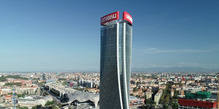 Un moderno grattacielo sinuoso, con l'insegna rossa "Generali" in cima, si erge contro un cielo azzurro e limpido, dominando un ampio paesaggio urbano con numerosi edifici e vegetazione.