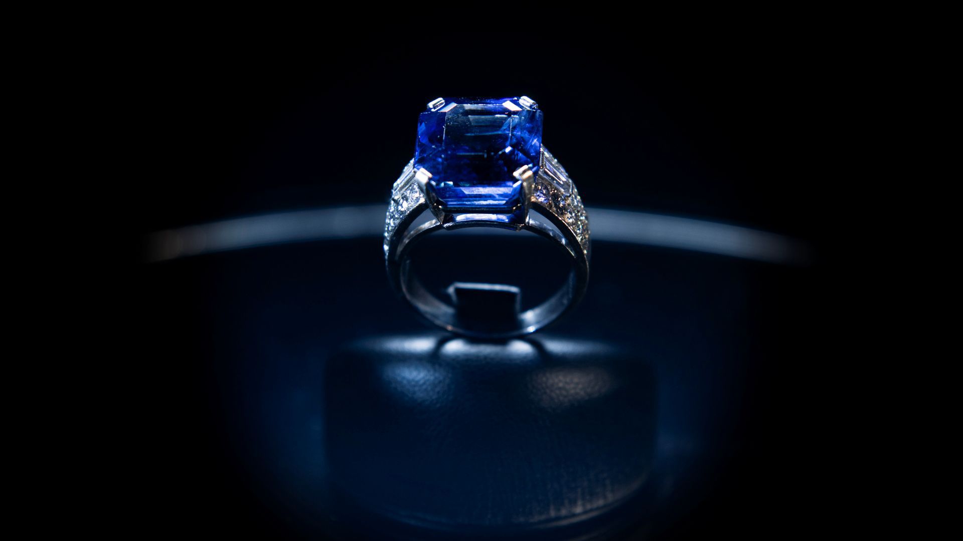 Un primo piano di un elegante anello con una grande pietra preziosa blu intenso incastonata in una fascia argentata riflettente. L'anello è esposto su un supporto nero su uno sfondo scuro e delicatamente illuminato, che ne evidenzia i dettagli intricati.