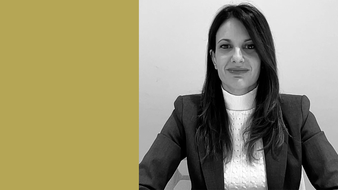 Valentina Guarise di BGR Tax & Legal, indossa un blazer e un maglione a collo alto è seduta a un tavolo e sorride. Lo sfondo è un muro semplice e un pannello giallo senape è sul lato sinistro dell'immagine. La foto è in bianco e nero.