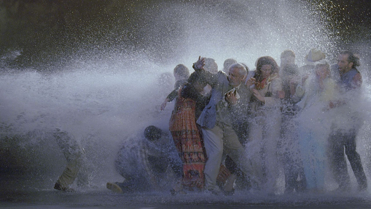 Un gruppo di persone, vestite con vari abiti, tra cui tailleur e abiti colorati, viene catturato da un'onda d'acqua. Ricordando un'installazione di Bill Viola, sembrano sorpresi e si riparano mentre l'acqua schizza intorno a loro.