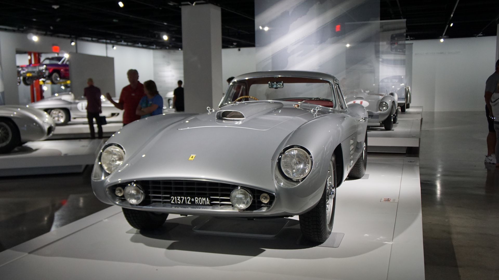 Un'auto sportiva d'epoca color argento è esposta in un moderno ambiente museale. L'auto è sollevata su una piattaforma bianca con una targa visibile. Altre auto simili sono disposte sullo sfondo e i visitatori sono visti osservare le esposizioni.