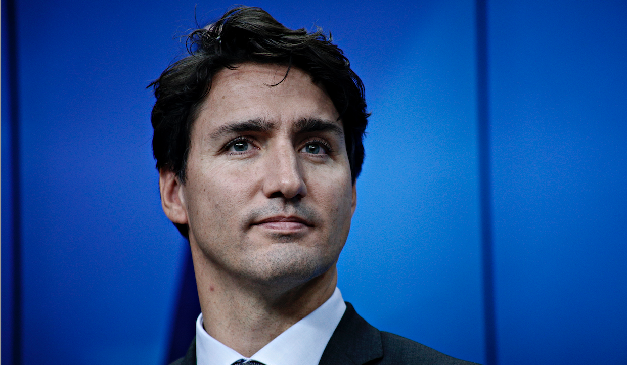 Justin Trudeau, presidente dimissionario del Canada, è in piedi su uno sfondo blu, guardando pensieroso in lontananza. Ha i capelli scuri e ondulati e un leggero sorriso sul viso.