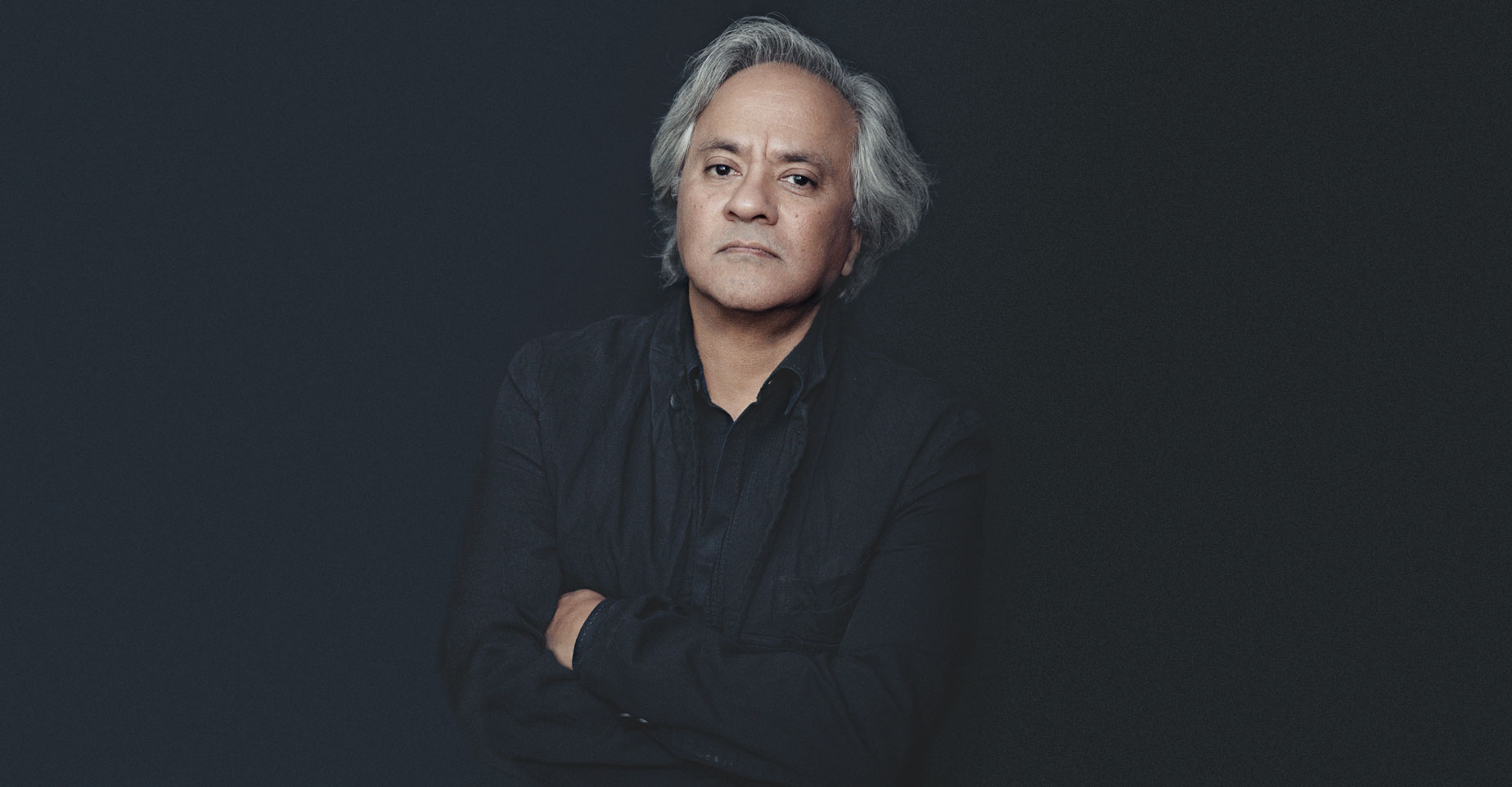 Una persona con i capelli grigi, che indossa una camicia scura che ricorda Anish Kapoor Nero, è in piedi su uno sfondo in ombra, con le braccia incrociate e lo sguardo leggermente rivolto verso la telecamera.