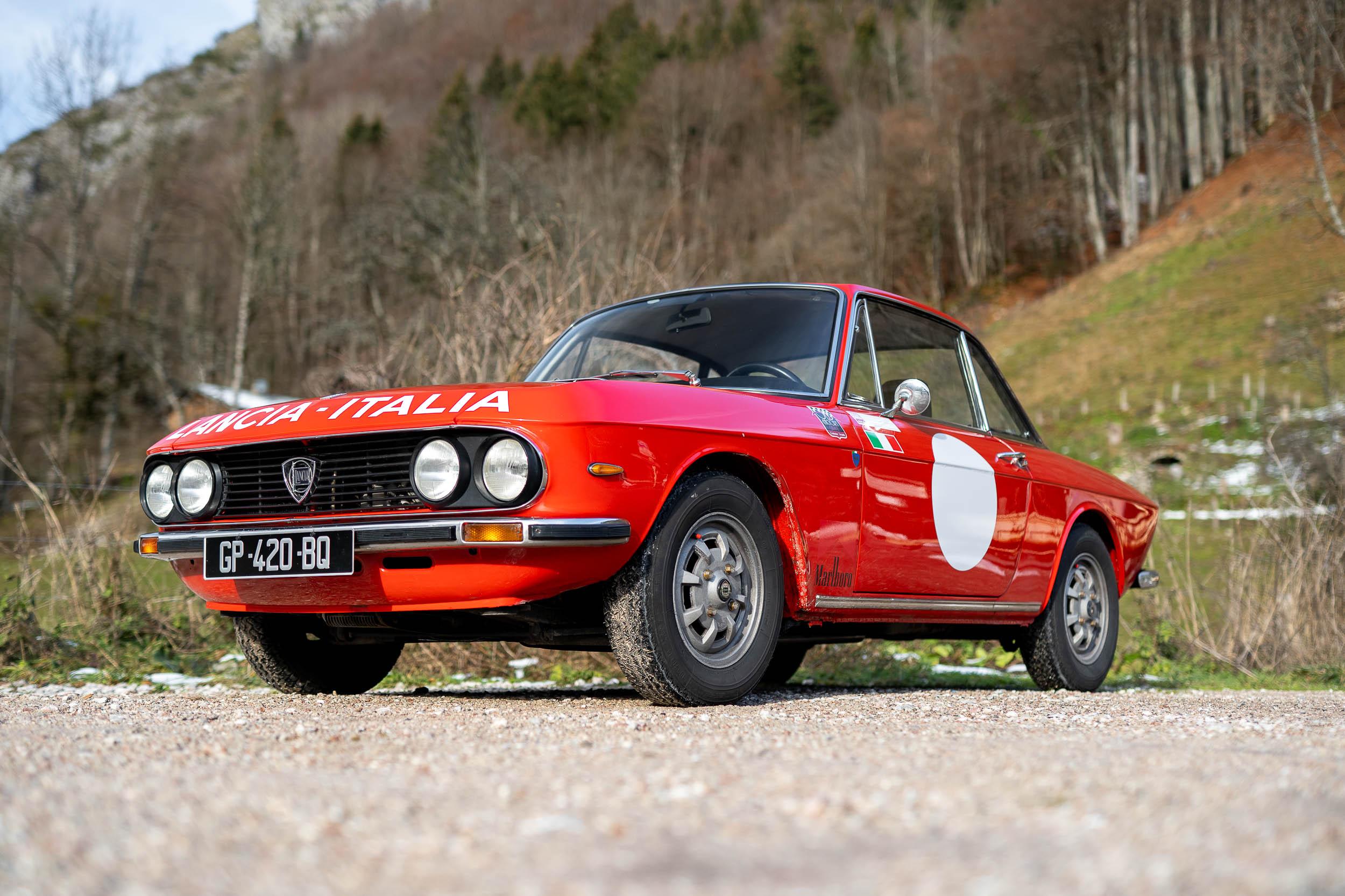Una classica Lancia Fulvia coupé rossa con un cerchio da corsa bianco sulla portiera è parcheggiata su una superficie ghiaiosa. L'auto presenta bandiere italiane sulla fiancata ed è posta su uno sfondo di alberi e colline.
