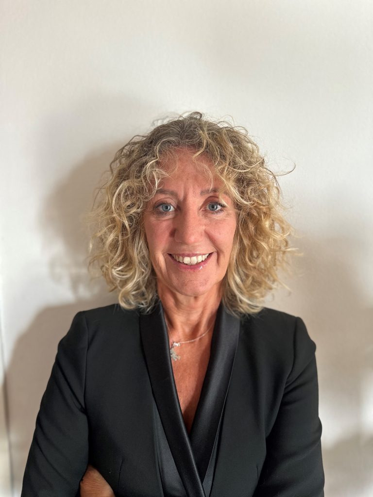Una donna sorridente con i capelli biondi e ricci, che indossa un blazer nero, è in piedi su uno sfondo bianco.