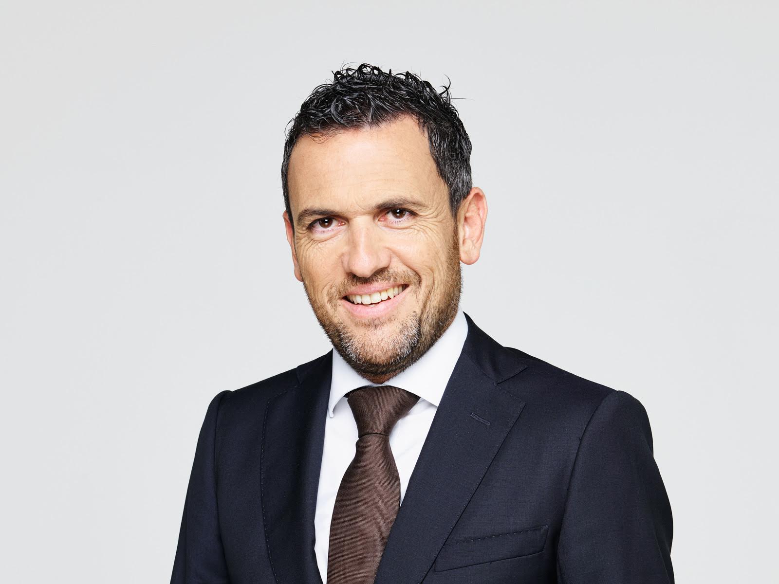 Uomo sorridente con capelli corti e ricci, vestito con un abito nero, una camicia bianca e una cravatta marrone scuro, in piedi su uno sfondo semplice e chiaro.