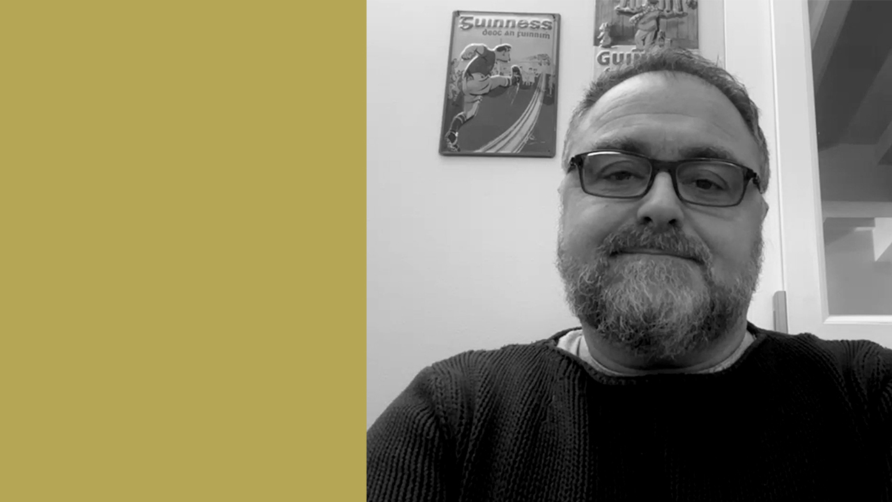 L'intervistato, Matteo Melodia, sorride leggermente mentre è seduta in un interno. Due poster incorniciati in stile vintage sono visibili sulla parete dietro di loro. L'immagine è in bianco e nero, con una fascia color senape sul lato sinistro.