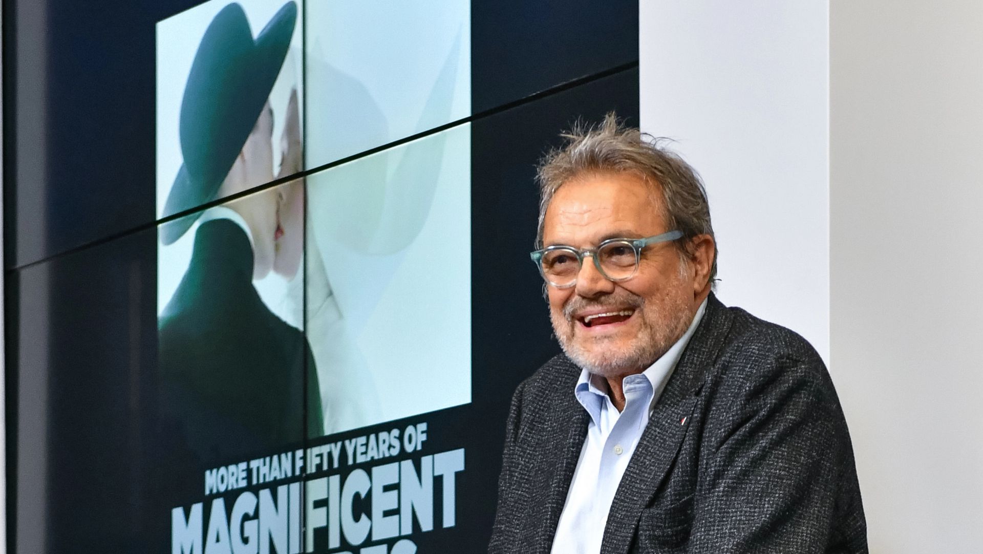 Un uomo sorridente con gli occhiali e la barba è seduto davanti a un manifesto con la scritta "Più di cinquant'anni di magnifici cappelli", che raffigura un primo piano di due persone che indossano grandi cappelli.