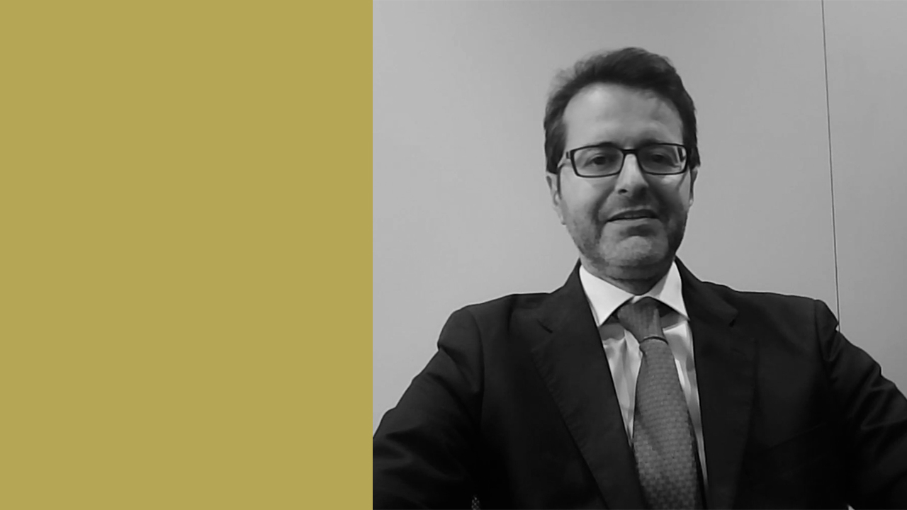 Marco Sandoli indossa occhiali e un completo con cravatta siede in un ambiente professionale. L'immagine è in bianco e nero, e mostra un contrasto tra la persona e uno sfondo semplice con una striscia gialla verticale sulla sinistra.