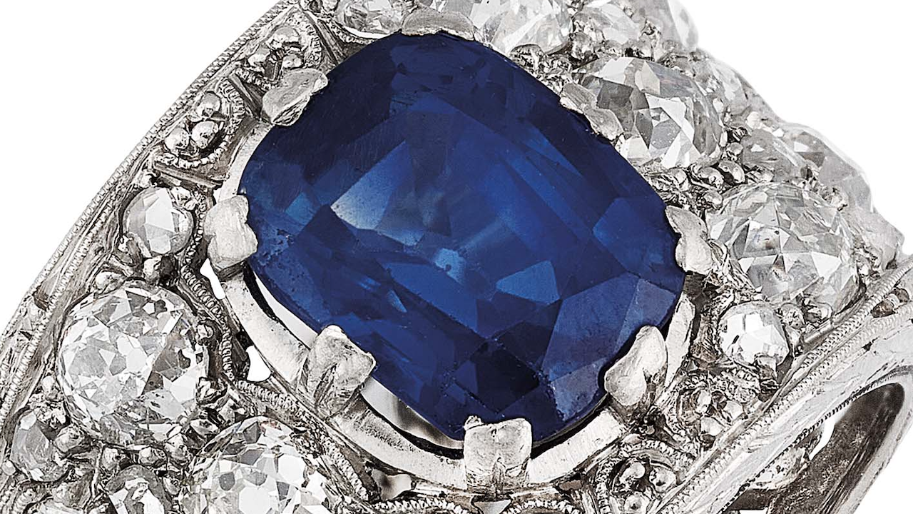 Primo piano di un anello d'argento con un grande zaffiro blu centrale circondato da diversi diamanti bianchi più piccoli in una montatura decorata.