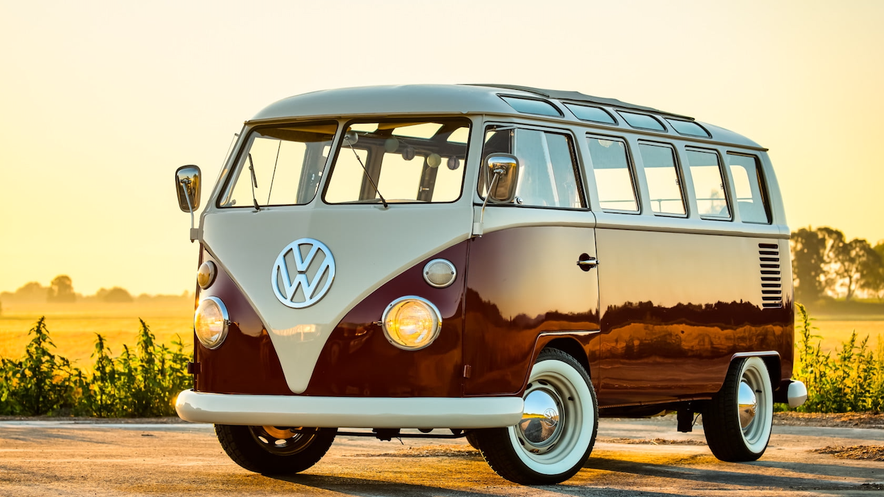 Un furgone Volkswagen Typ 2 d'epoca parcheggiato su una strada al tramonto. Il veicolo è bicolore, con vernice color crema e bordeaux. La luce del sole proietta un caldo chiarore, evidenziando l'iconico emblema VW sulla parte anteriore. Lo sfondo mostra un campo e degli alberi.