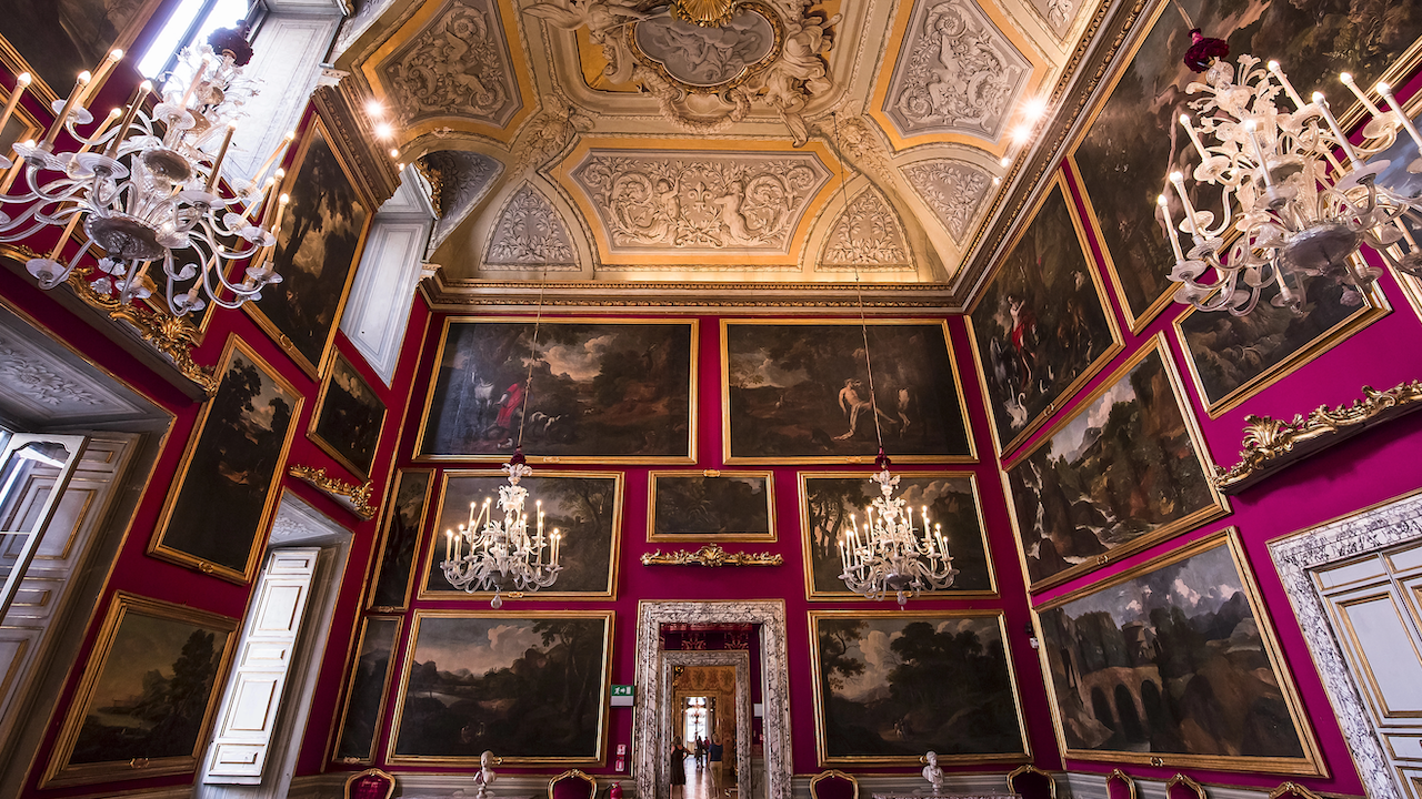 Una sala sontuosa adornata da lampadari decorati e numerosi grandi dipinti in cornici dorate su pareti rosso scuro. Il soffitto finemente decorato presenta eleganti intagli e modanature. Un grande portone è aperto al centro della sala.