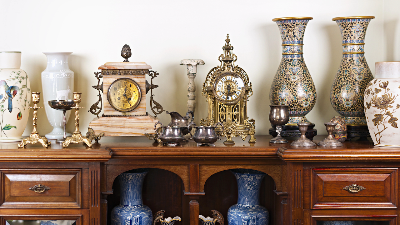 Un'esposizione di decorazioni vintage su una credenza in legno, con due orologi decorati, grandi vasi in ceramica con motivi intricati, portacandele in argento e ottone e diversi pezzi decorativi. La credenza ha vasi blu e bianchi all'interno.