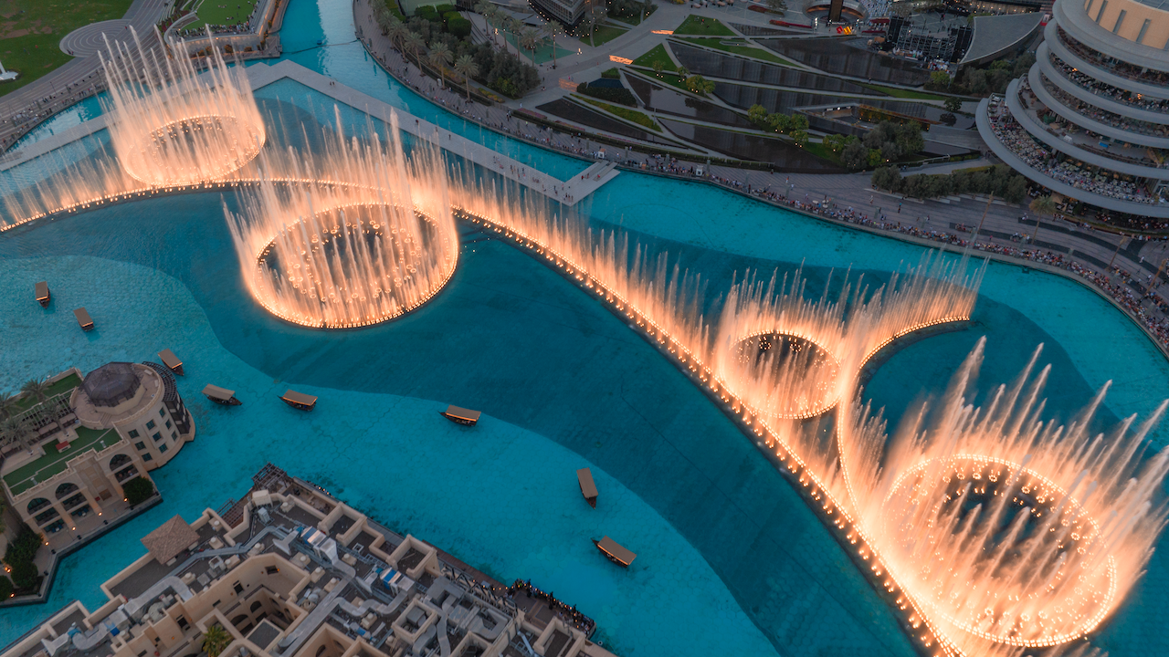 Vista aerea di Dubai con una grande fontana con motivi circolari e getti d'acqua illuminati in una grande piscina. Edifici e passerelle circondano la piscina e piccole imbarcazioni sono visibili sull'acqua.