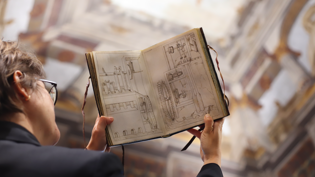 Una persona che indossa occhiali tiene in mano un album da disegno aperto con intricati disegni meccanici. Lo sfondo mostra un soffitto sfocato e decorato, che suggerisce un ambiente architettonico o storico.