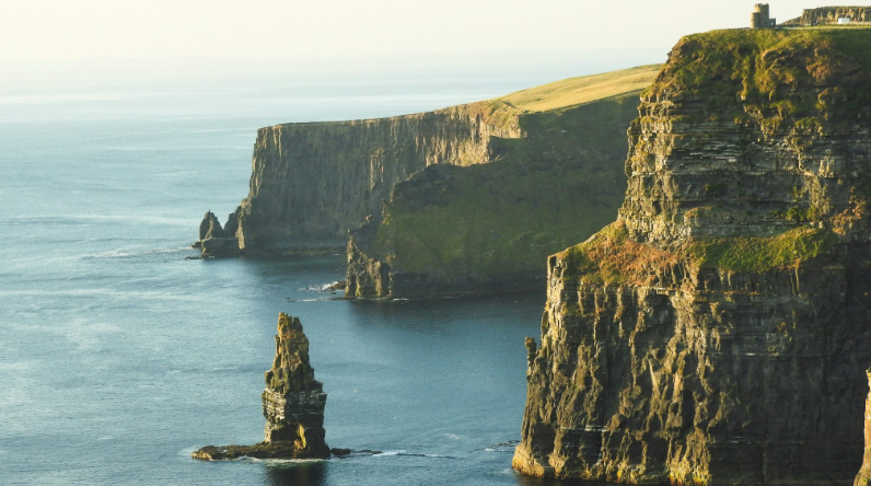 Cliffs overlooking the ocean on a clear day with green grass on top, stone formations jutting from the water, and a structure perched on an edge. The scene captures dramatic vertical cliffs against a serene blue sea.