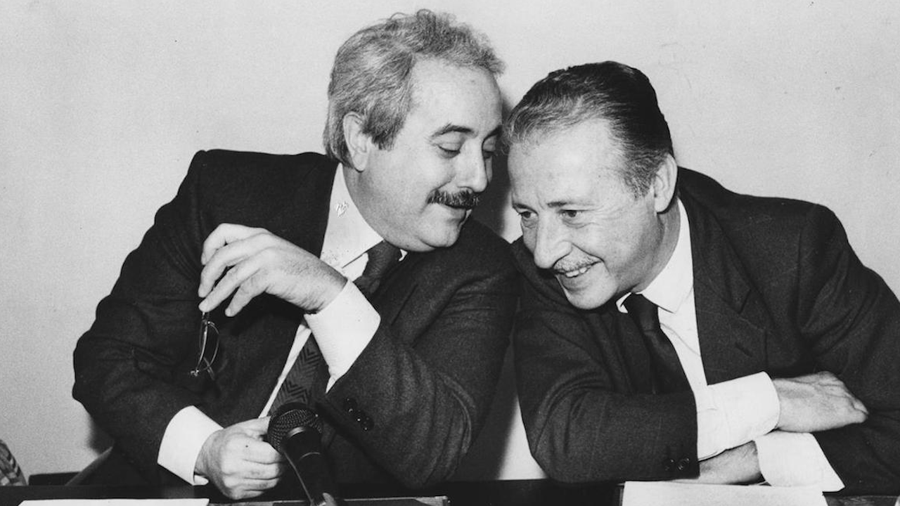 Falcone e Borsellino condividono un momento di leggerezza a un tavolo. Uno si sporge verso l'altro con un sorriso, tenendo gli occhiali in una mano. Entrambi sembrano impegnati in una conversazione amichevole. Un microfono è posizionato sul tavolo di fronte a loro.