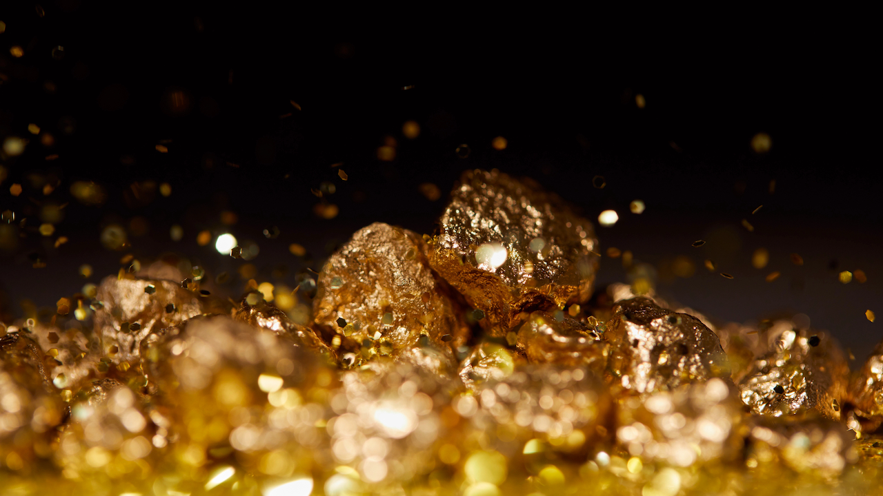 Primo piano di pepite d'oro luccicanti sparse su uno sfondo scuro. La ricca consistenza metallica e il caldo bagliore delle pepite sono evidenziati, con alcune particelle d'oro più piccole attorno a loro.