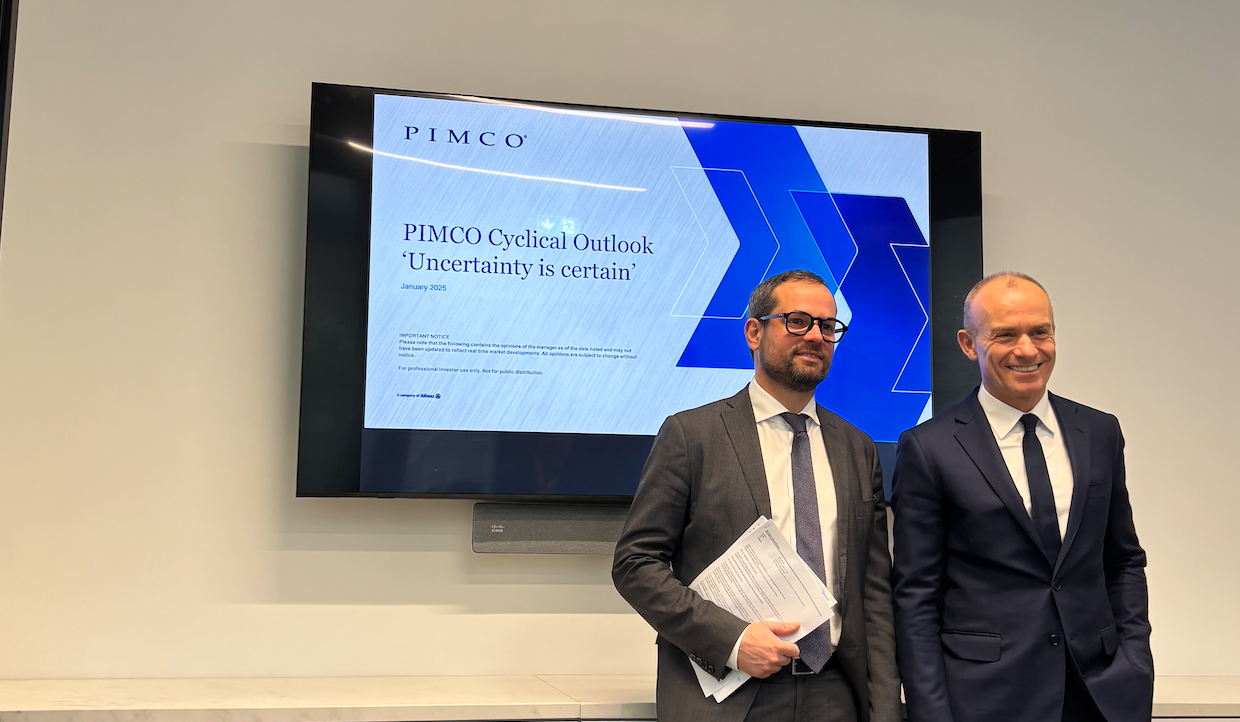 Due uomini in giacca e cravatta stanno sorridendo davanti a una diapositiva di presentazione. La diapositiva, intitolata "PIMCO Cyclical Outlook 'Uncertainty is certain'", è visualizzata su un grande schermo dietro di loro. Un uomo tiene in mano dei documenti ed entrambi sembrano professionali e coinvolti.