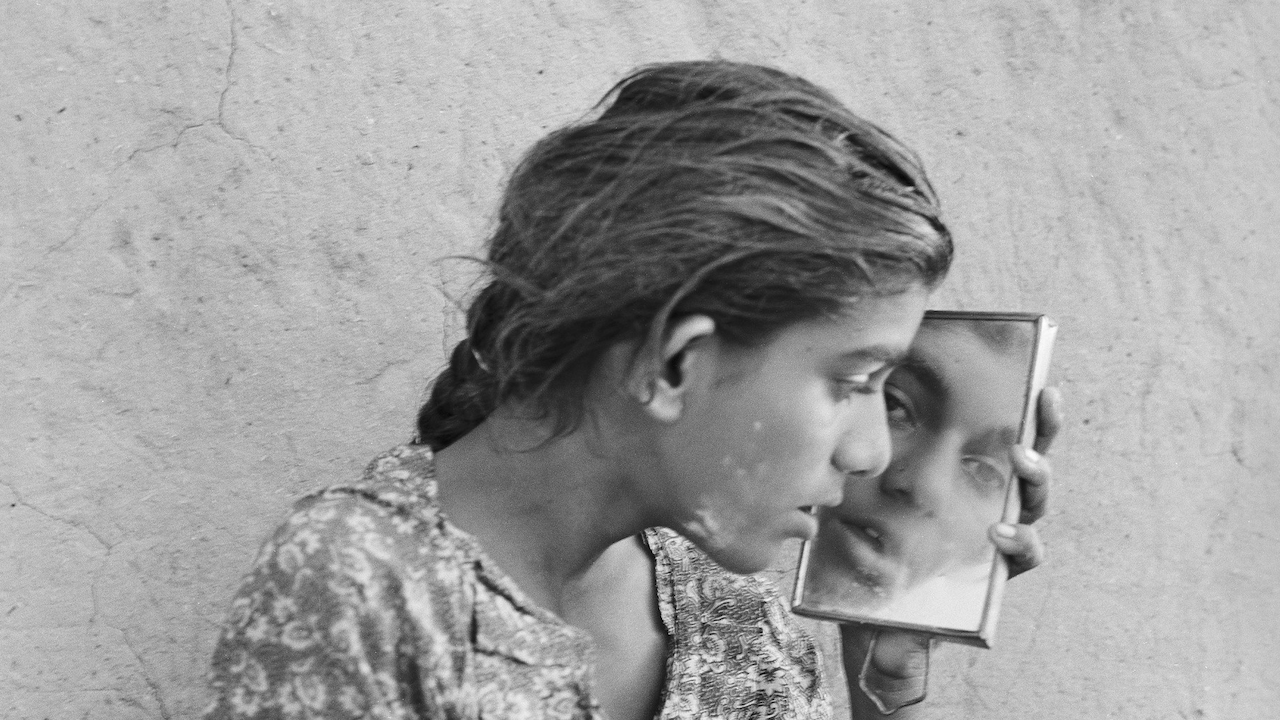 Un'immagine in bianco e nero di un giovane che tiene un piccolo specchio vicino al viso, riflettendo la sua immagine. Ha i capelli intrecciati e indossa una camicia fantasia, seduto contro una parete ruvida. L'espressione sembra contemplativa.
